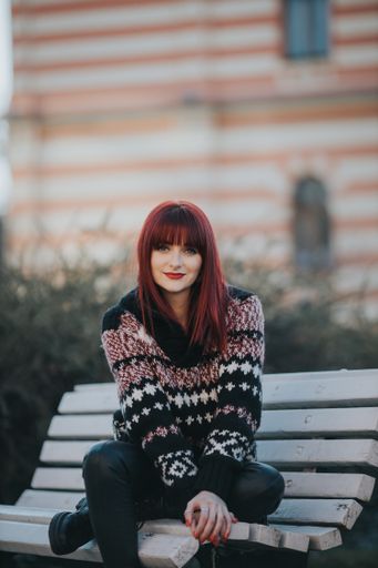 A vertical shot of a young smiling female with red hair posing in a warm sweater sitting on a bench