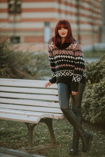 A vertical shot of a young caucasian female with red hair in a cozy sweater standing near a bench