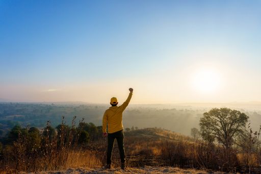 A back view of a young male wearing a yellow hoodie and cap with his fist up in a field at sunset