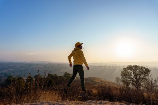 A young Mexican male wearing a yellow hoodie and cap jumping up in a field against a sunset sky