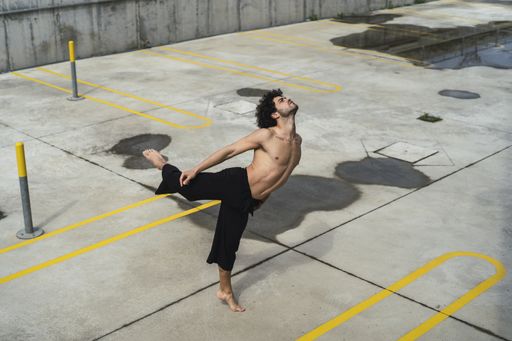 A Caucasian curly-haired topless man dancing in a park on a sunny day in Spain