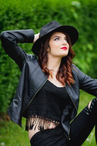 A portrait of a confident stylish female wearing a leather jacket paired with a fedora hat in a park