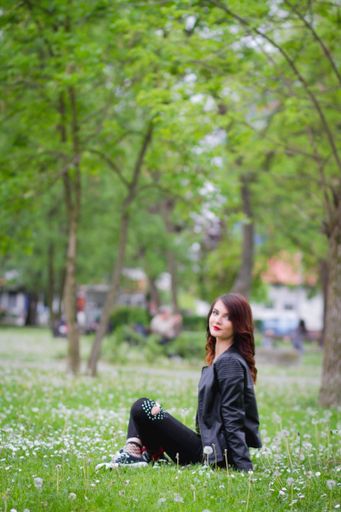 A shallow focus of a young stylish female wearing jeans with a leather jacket and sitting on the ground in a park
