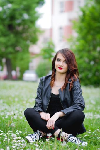 A shallow focus of a young stylish female wearing jeans with a leather jacket and sitting on the ground in a park