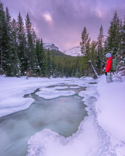 A hiker by a frozen river in snowy mountains looking at the purple sunset sky