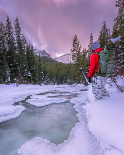 A vertical shot of hiker by a frozen river in snowy mountains looking at the purple sunset sky