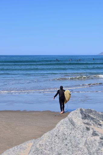 A vertical shot of a surfer walking out to the ocean with a large boulder in the foreground up close