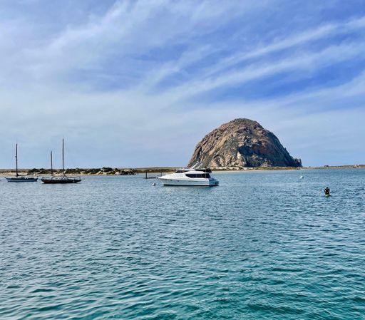 A white Yacht on the water in front of Morro Bay Rock with two smaller boats and a Surfer on a sunny day