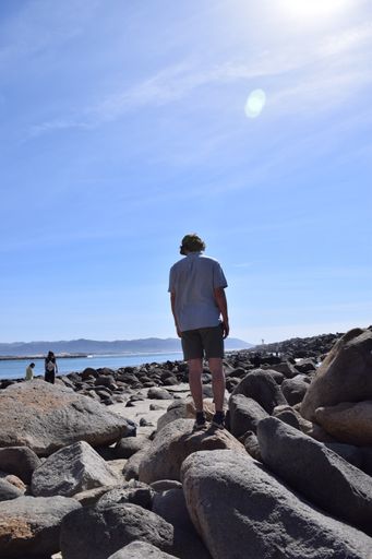 A photo of a mans back facing the water as he stands on boulders looking out in casual clothing on a beautiful warm sunny day with blue sky background
