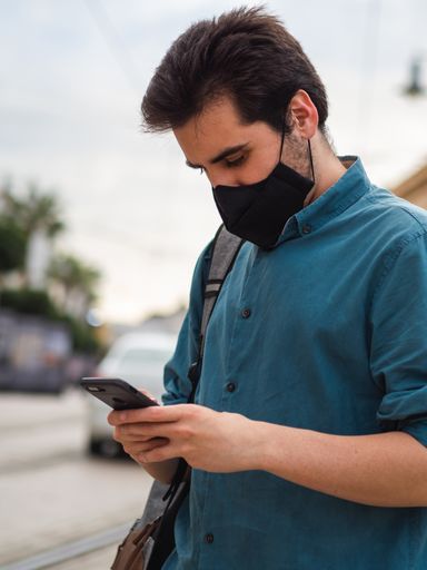 A vertical shot of Spanish male with face mask using phone outdoors - concept of the new normal