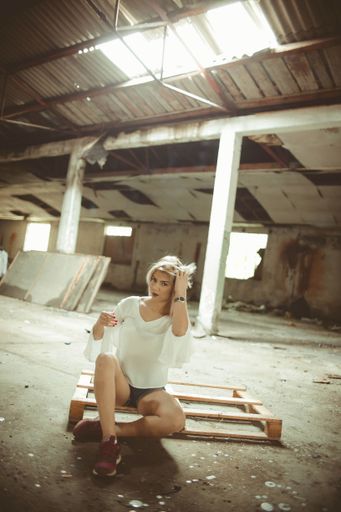 A vertical shot of a young blonde female posing sitting on a wooden plank inside an abandoned ruined building