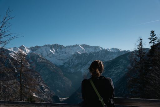 An adult female stands on a rocky outcrop with a breathtaking view of a picturesque snowy mountain range and evergreen trees in the background