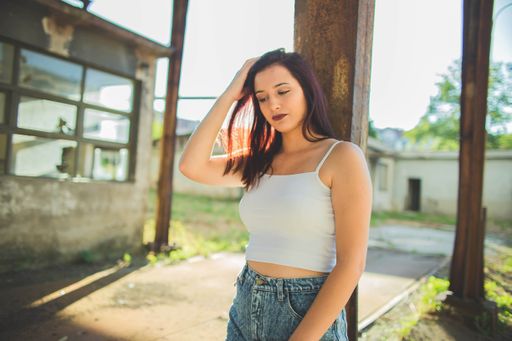 A shallow focus shot of a Caucasian woman leaning on a rusty pole, looking down, and playing with her hair surrounded by abandoned buildings