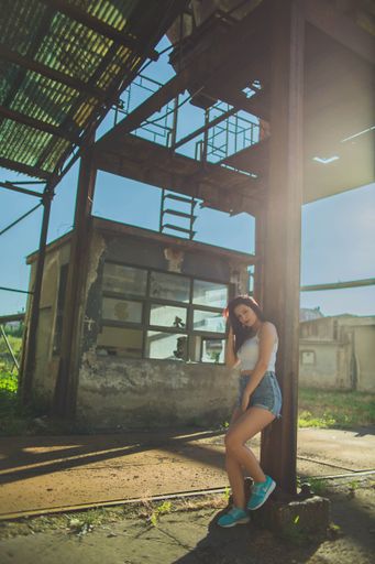 A vertical shot of a Caucasian girl in shorts leaning against metal structure, looking at the camera while playing with her hair