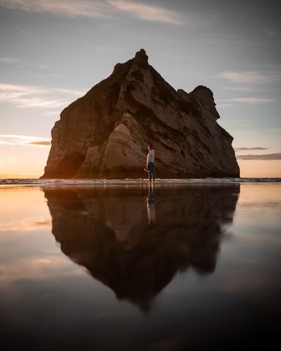 A vertical shot of a person standing on a cliff reflecting on the sea during the sunset