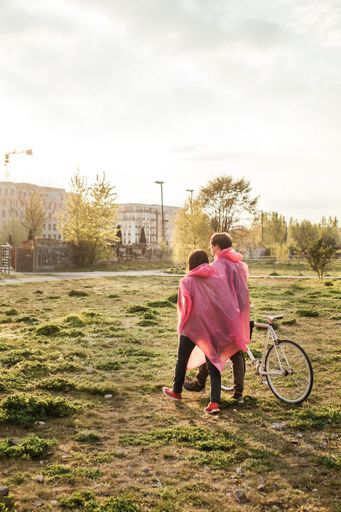 A vertical shot of a male and a female in pink plastic raincoats walking in the field with a bicycle on a date
