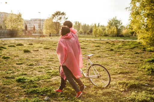 A male and a female in pink plastic raincoats walking in the field with a bicycle on a date