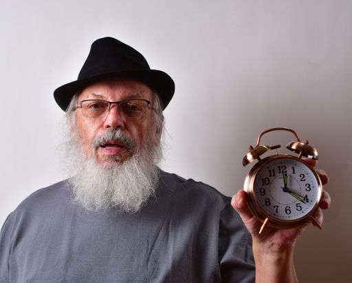 An old American male in a black hat and eyeglasses holding an alarm clock