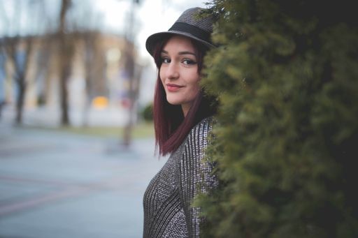 A Caucasian woman in a gray hat with red hair standing in front of a bush, smiling at the camera