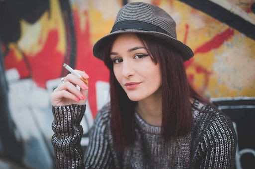 A young Caucasian woman in a hat standing in front of graffiti wall, looking at the camera with a cigarette in her hand