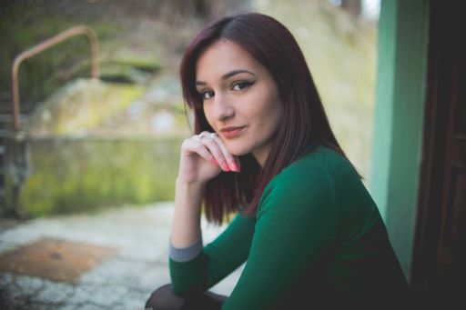 A young Caucasian woman looking at the camera in front of house with her hand on her chin and sun shining behind her
