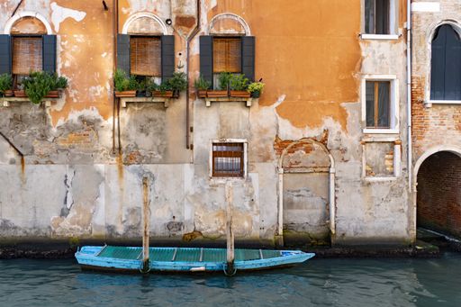 An old orange building with open windows and plants and a blue boat in the Venice canal, Italy