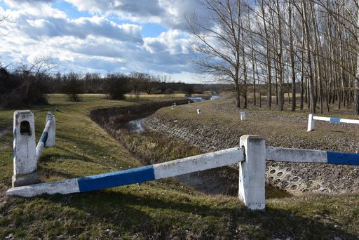 A river flowing amid field with dry trees under the cloudy sky on the foreground of a white-blue fence