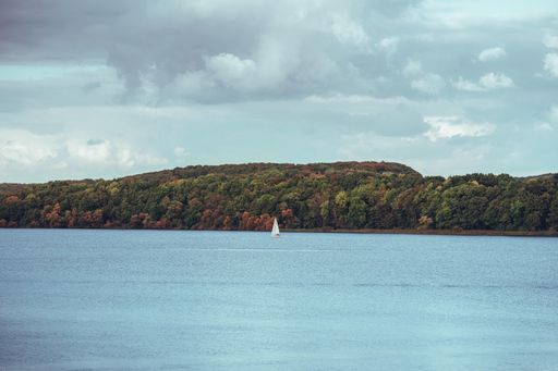 A beautiful shot of a white sailboat on a sea with green mountains in the background under a cloudy sky