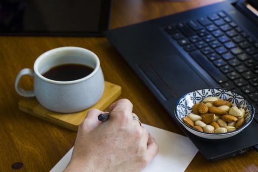 A person writing in a notebook while working on a laptop and drinking coffee