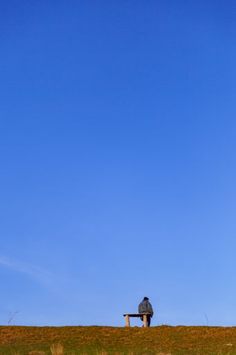 A vertical back view of a person sitting on a bench with the blue sky in the background
