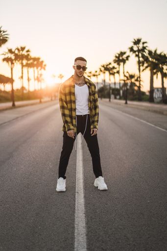 A vertical shot of a handsome Spanish Caucasian man with tattoos and sunglasses posing outdoors in the middle of the street during sunset
