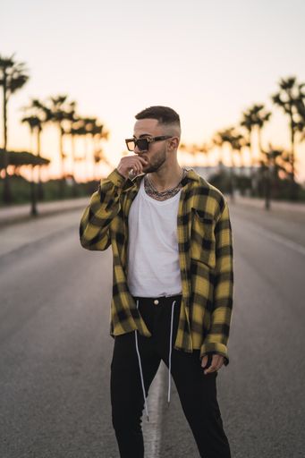 A vertical shot of a handsome Spanish Caucasian man with tattoos and sunglasses posing outdoors in the middle of the street during sunset