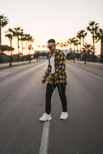 A vertical shot of a handsome Spanish Caucasian man with tattoos and sunglasses posing outdoors in the middle of the street during sunset