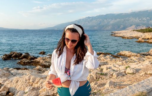 A young Caucasian female in a swimsuit and sunglasses posing with a piece of watermelon at the beach
