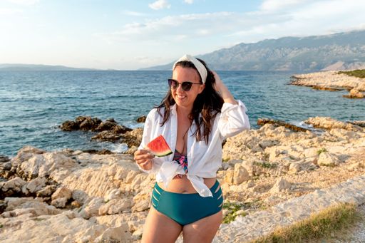A young Caucasian female in a swimsuit and sunglasses posing with a piece of watermelon at the beach