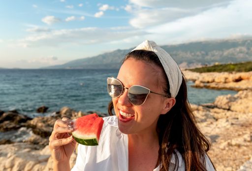 A young Caucasian female in a white shirt and sunglasses eating a piece of watermelon at the beach