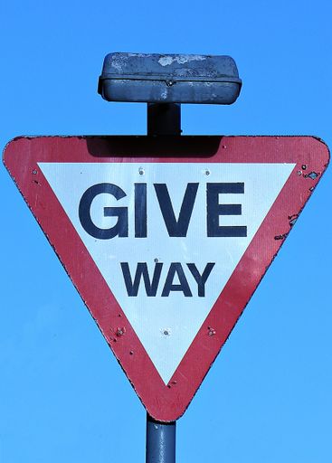 Weathered street sign, inverted triangle shape, on which words "GIVE WAY" are printed in black capital letters on white background. Red border around