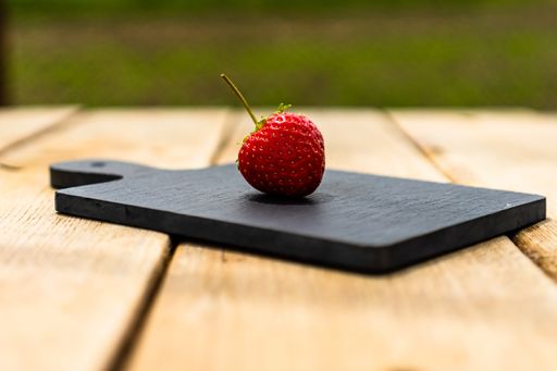 A closeup of one ripe strawberry on small black cutting board placed outdoor on a wooden table