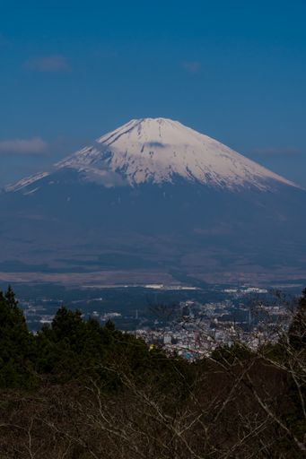 A bird's-eye vertical view of snow-capped Mount Fuji through clouds and trees, with a city at its feet, on a sunny spring day in Japan