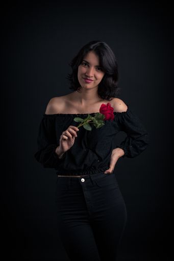 A portrait shot of a beautiful Hispanic woman smiling on the camera holding a red rose in front of a plain black background.