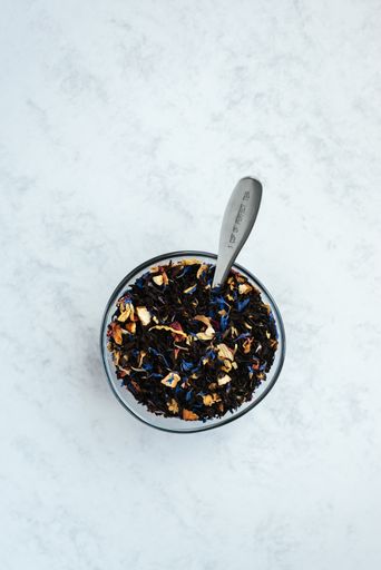 A vertical shot of a bowl and spoon full of black tea mixed with flower petals on a light background.