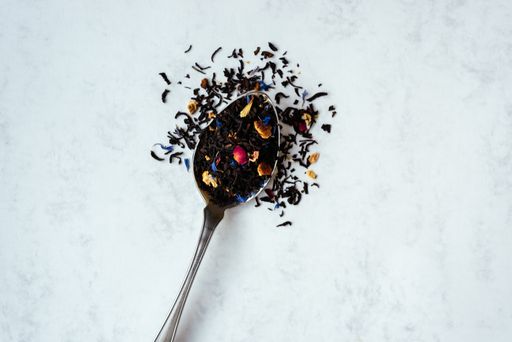 A top view close-up shot of a spoon full of black tea mixed with flower petals on a light background with copy space.