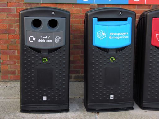 Two recycling bins or cans standing next to each other before a brick wall. One is for food and drink cans, the other is for newspapers and magazines.