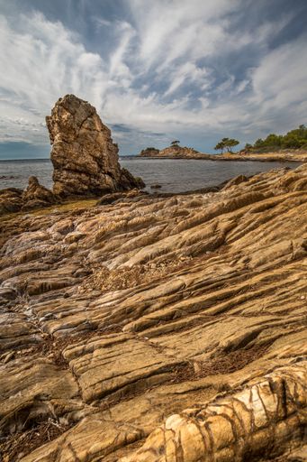 A vertical picture of rocks surrounded by the sea and greenery under the cloudy sky and sunlight