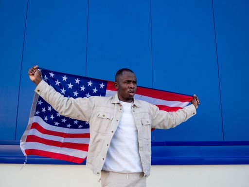 A Black Spanish male with hands up holds a flag of the USA behind him against a blue wall