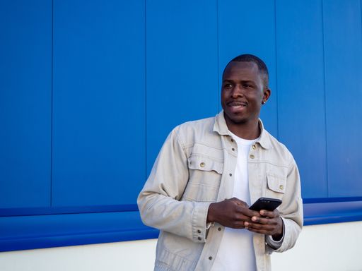 A fashionable black Spanish male using his smartphone against a blue wall