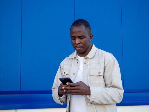 A fashionable black Spanish male using his smartphone against a blue wall