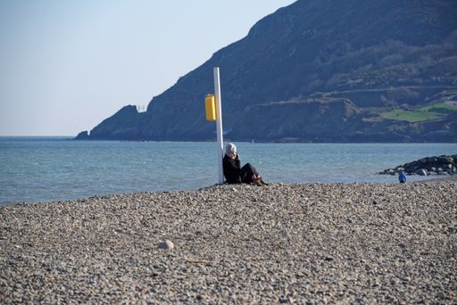 A woman enjoys the weather as she sits leaning against the lifebuoy post on pebble beach in Bray, Co. Wicklow, Ireland on sunny spring day.