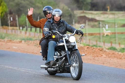 two men on motorcycles with protective gear riding on tar road waving greeting