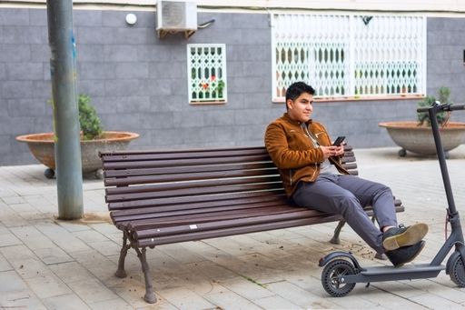 A young Latin guy in a brown jacket resting on a park bench after a ride on the electric scooter